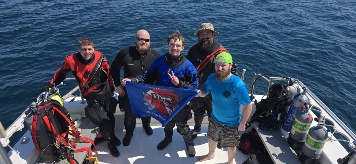 Five Deep Adventures divers on a dive boat holding the Deep Adventures flag, open water behind them